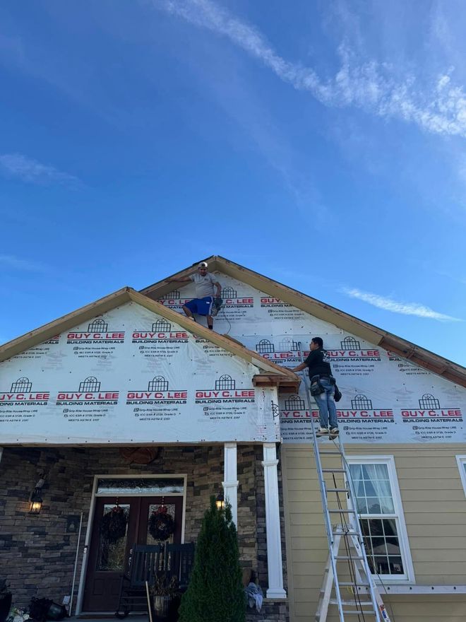 Two workers on ladders install house wrap on the exterior wall of a building under a bright blue sky.