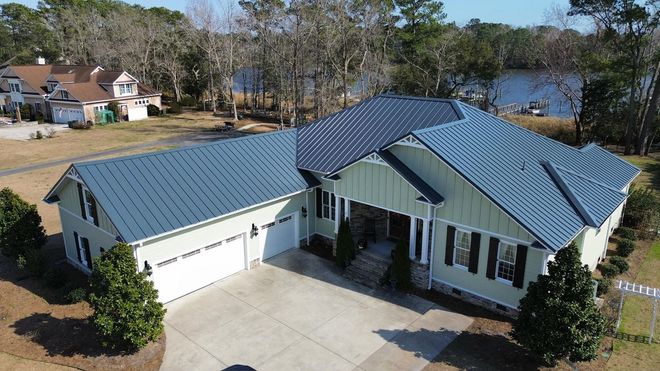 Aerial view of a pale green house with a dark blue metal roof, white garage doors, and nearby trees by a river.