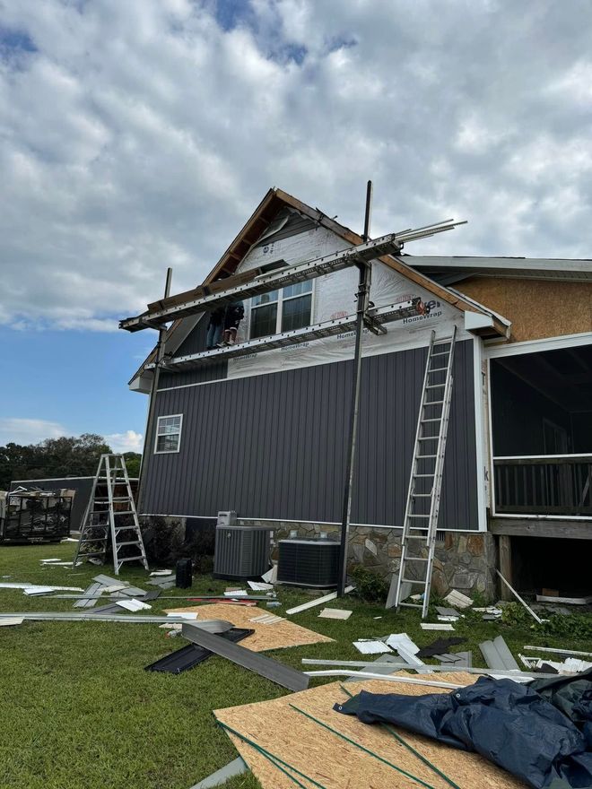 A side view of a house undergoing construction with dark gray siding, scaffolding, ladders, and debris on the ground.