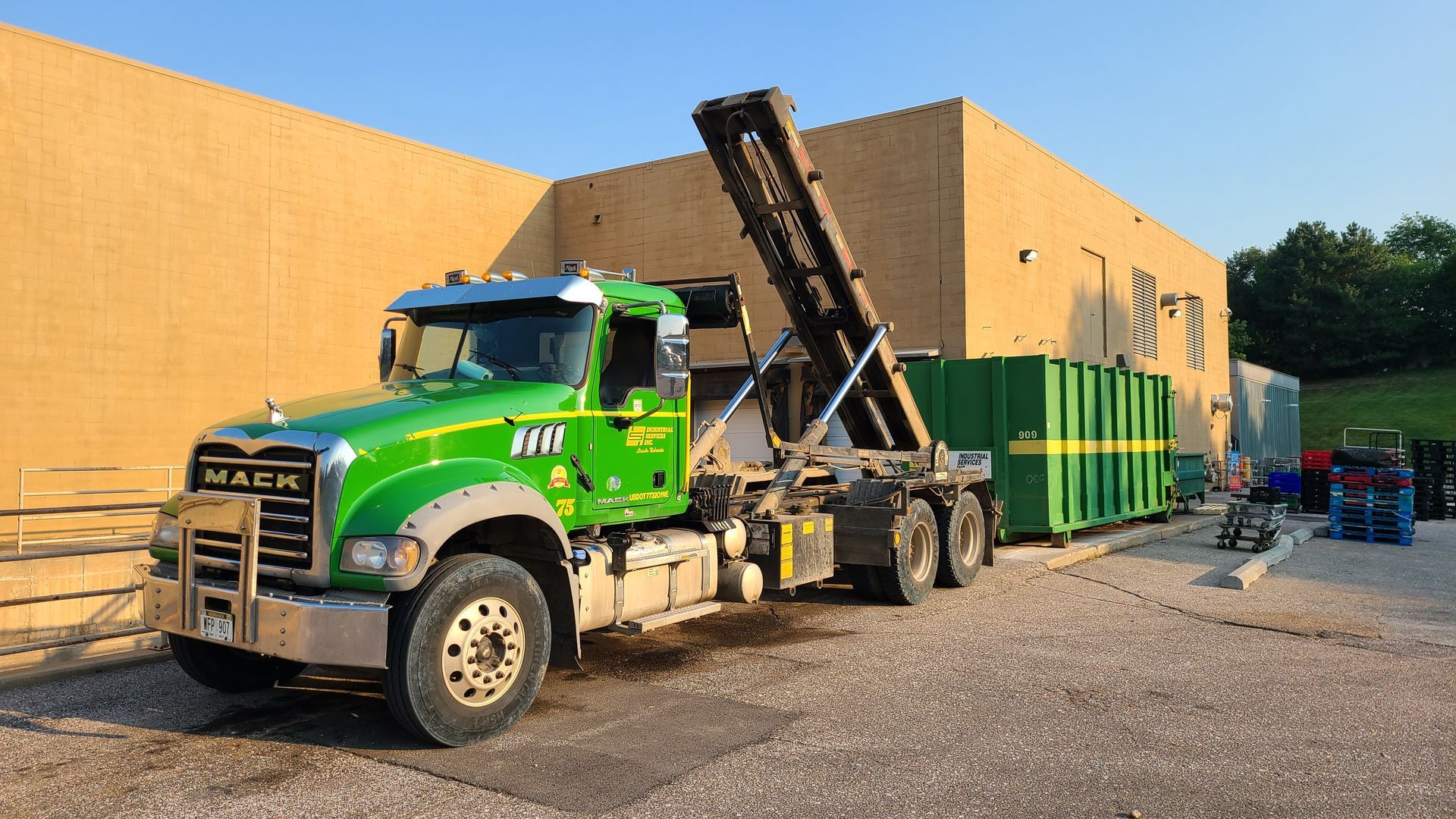 Green Mack truck lifting a green container beside a beige building. Green Mack truck lifting a green container beside a beige building.