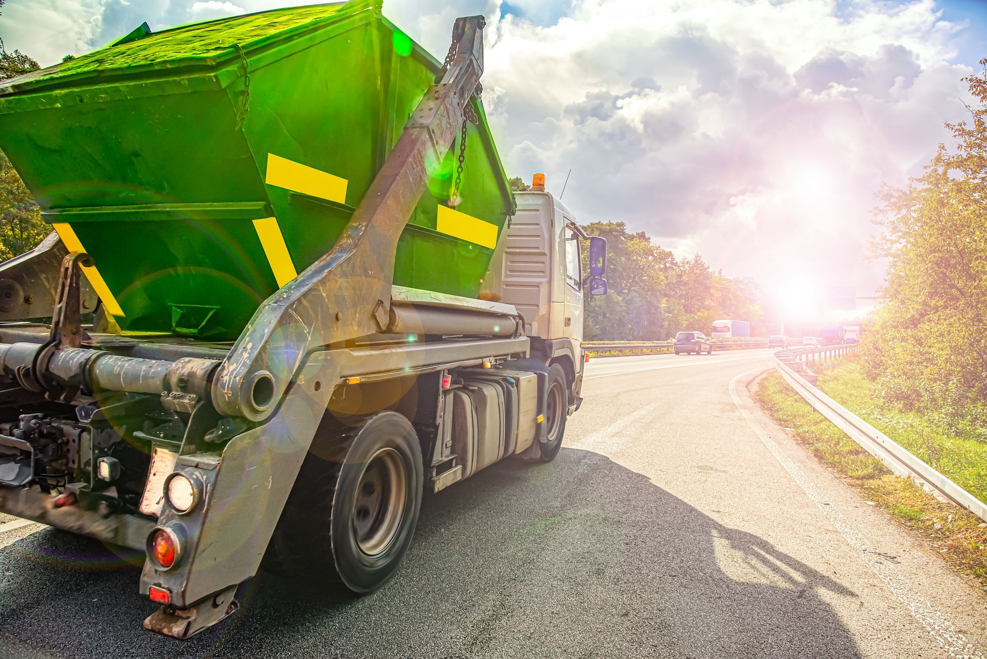 Garbage truck transporting a large green waste container along a sunlit road.