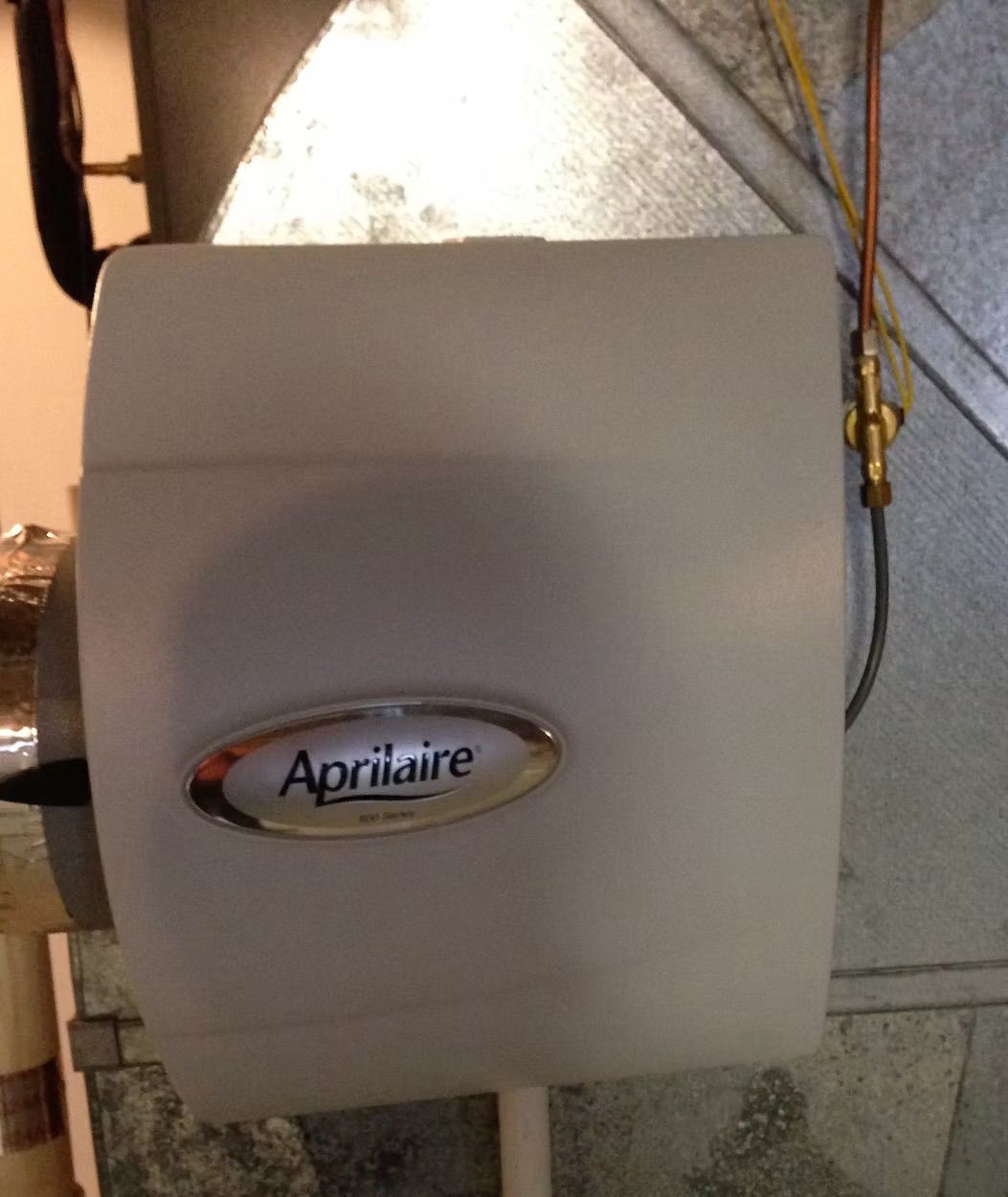 Aprilaire humidifier mounted on ductwork, gray with oval logo and tubing.