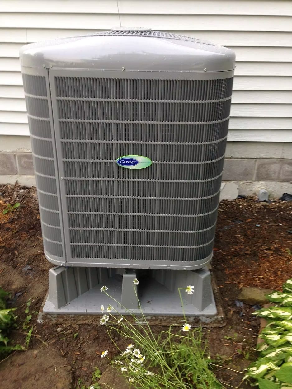 Gray Carrier air conditioning unit on a concrete base next to a white house and dirt with greenery.