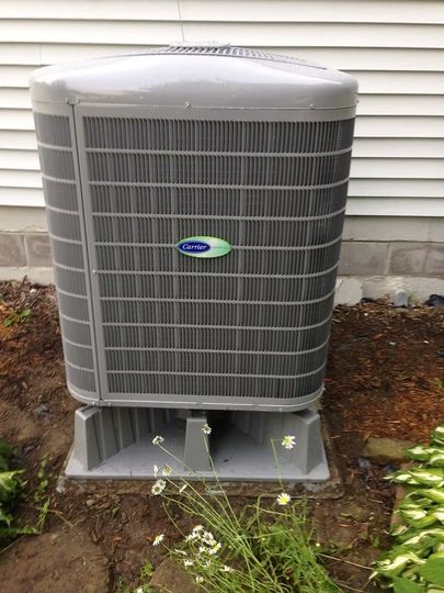 Gray Carrier air conditioning unit on a concrete base next to a white house and dirt with greenery.