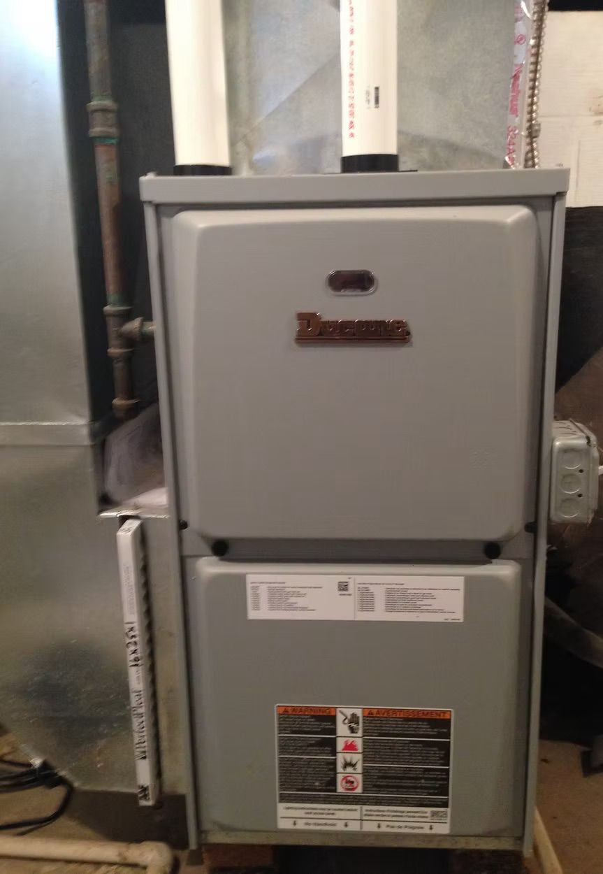 Gray furnace with white pipes, a brass nameplate, and warning labels in a basement.