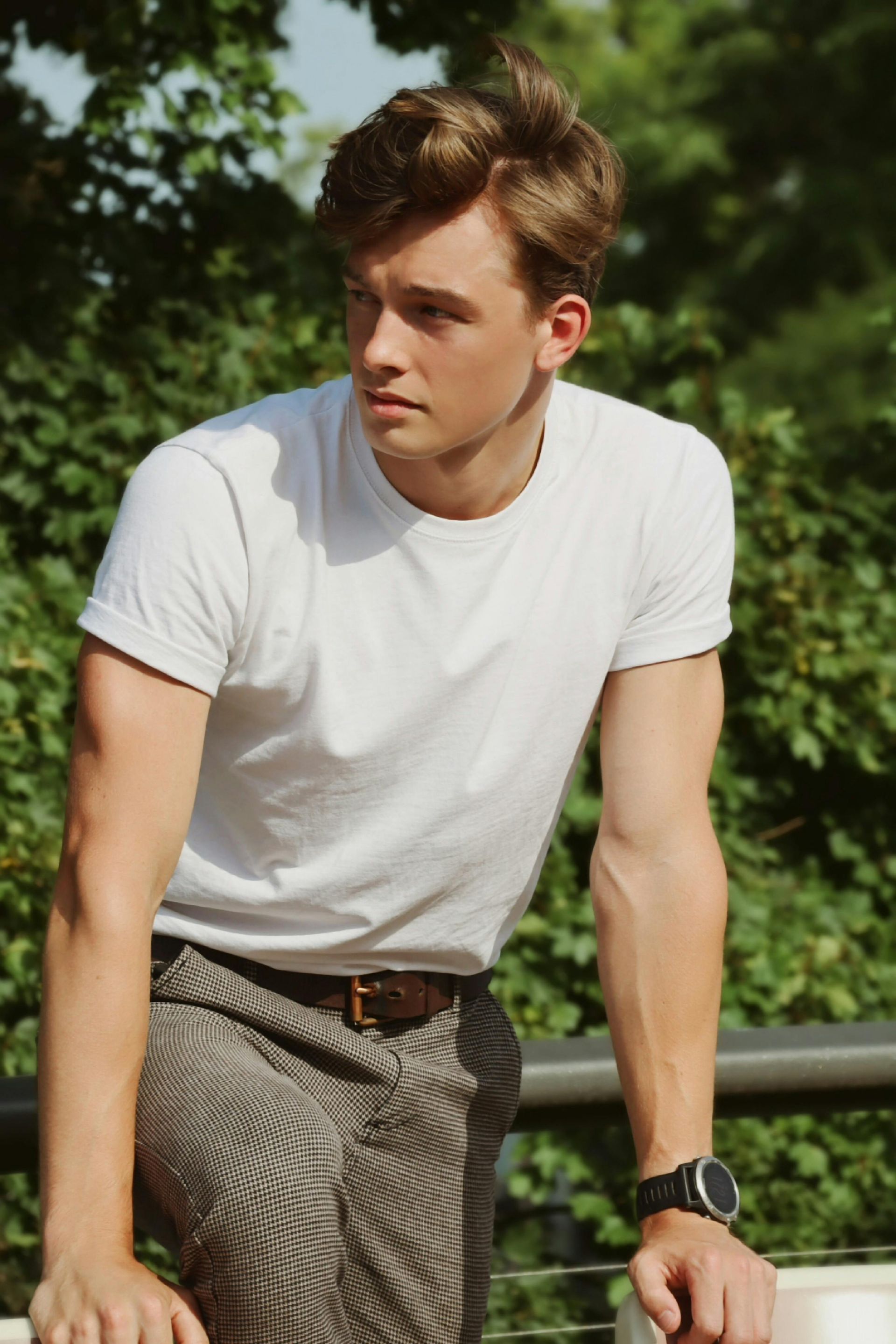 Man in white t-shirt and patterned pants leaning on a railing, looking to the side. Outdoors.