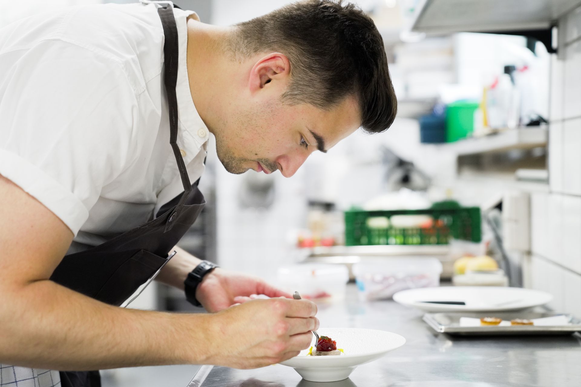 En person i hvid uniform og forklæde pynter omhyggeligt en ret i et professionelt køkken.