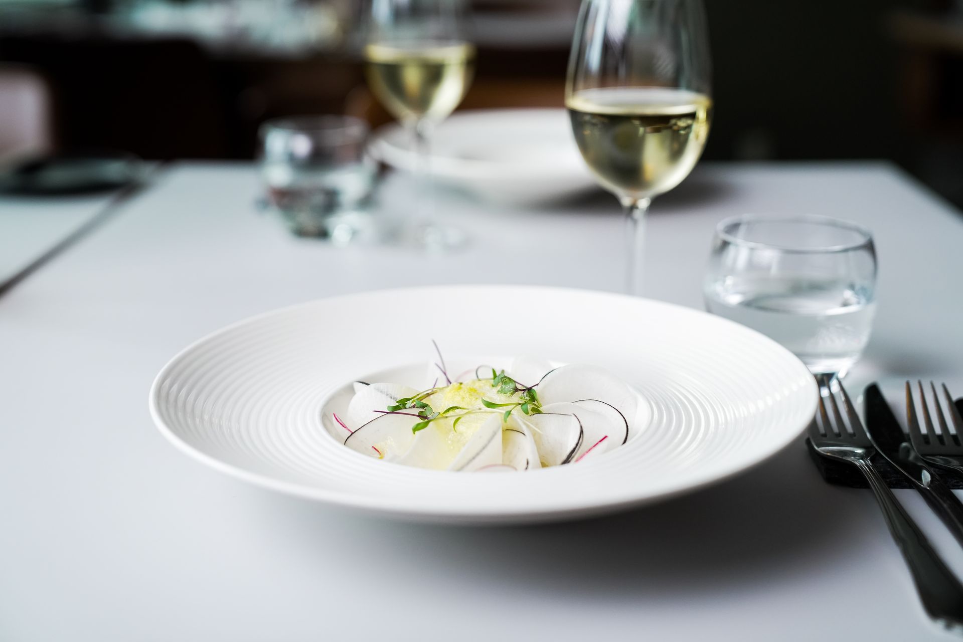 En gourmetforret bestående af tyndt skåret hvid fisk serveret i en lav, tekstureret hvid skål på et formelt spisebord.