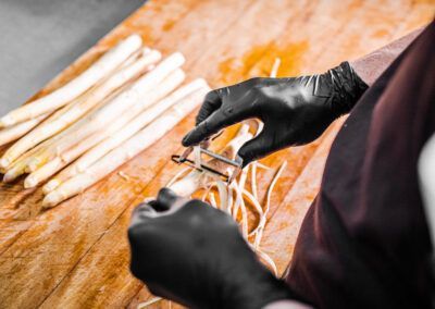 Person wearing black gloves peeling white asparagus on a wooden surface.