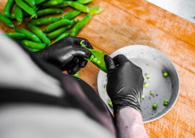 Person wearing black gloves shelling fresh green peas into a white bowl on a wooden surface.