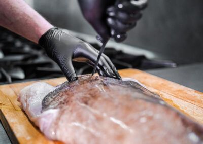 Person in black gloves filleting a fish on a wooden board with a sharp knife.