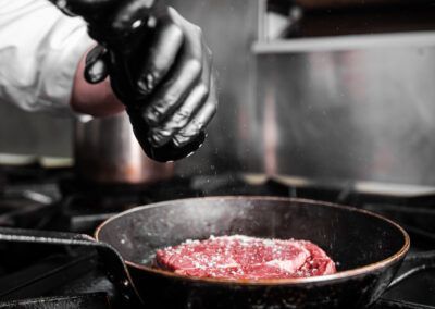 Chef seasoning a steak with salt in a hot pan, in a kitchen setting.