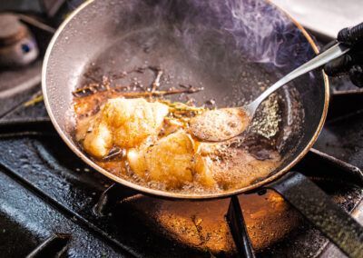 Fish fillets searing in a pan with bubbling butter and herbs over a gas stove.
