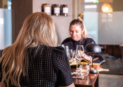 Two women at a restaurant table. One with long blonde hair, other sampling wine. Interior setting.