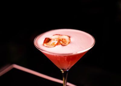 Pink cocktail with foamy top in a martini glass, garnished with a dried fruit slice, against a black background.