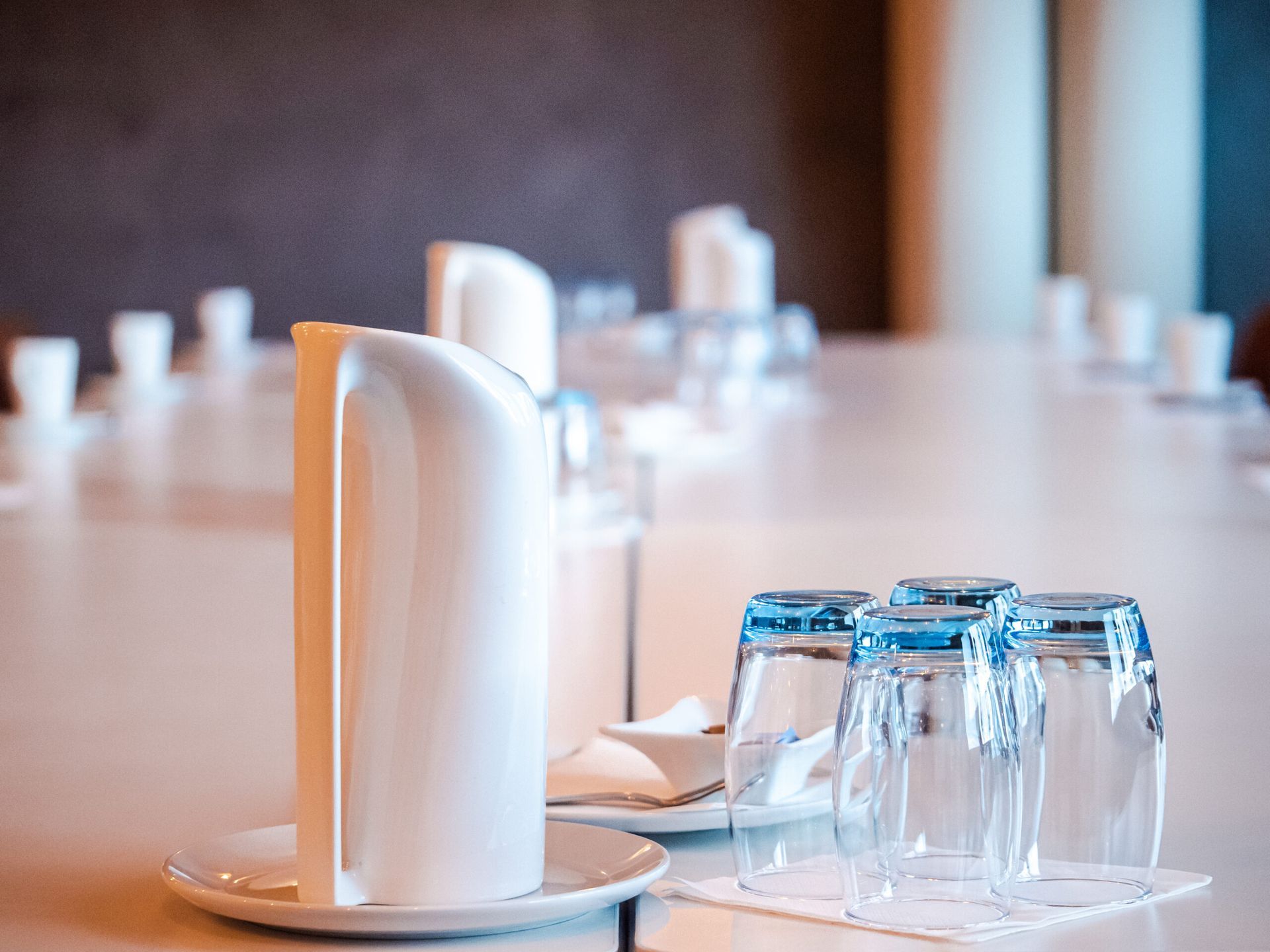 A white conference table set with glasses, napkins, and plates in a room with dark walls.