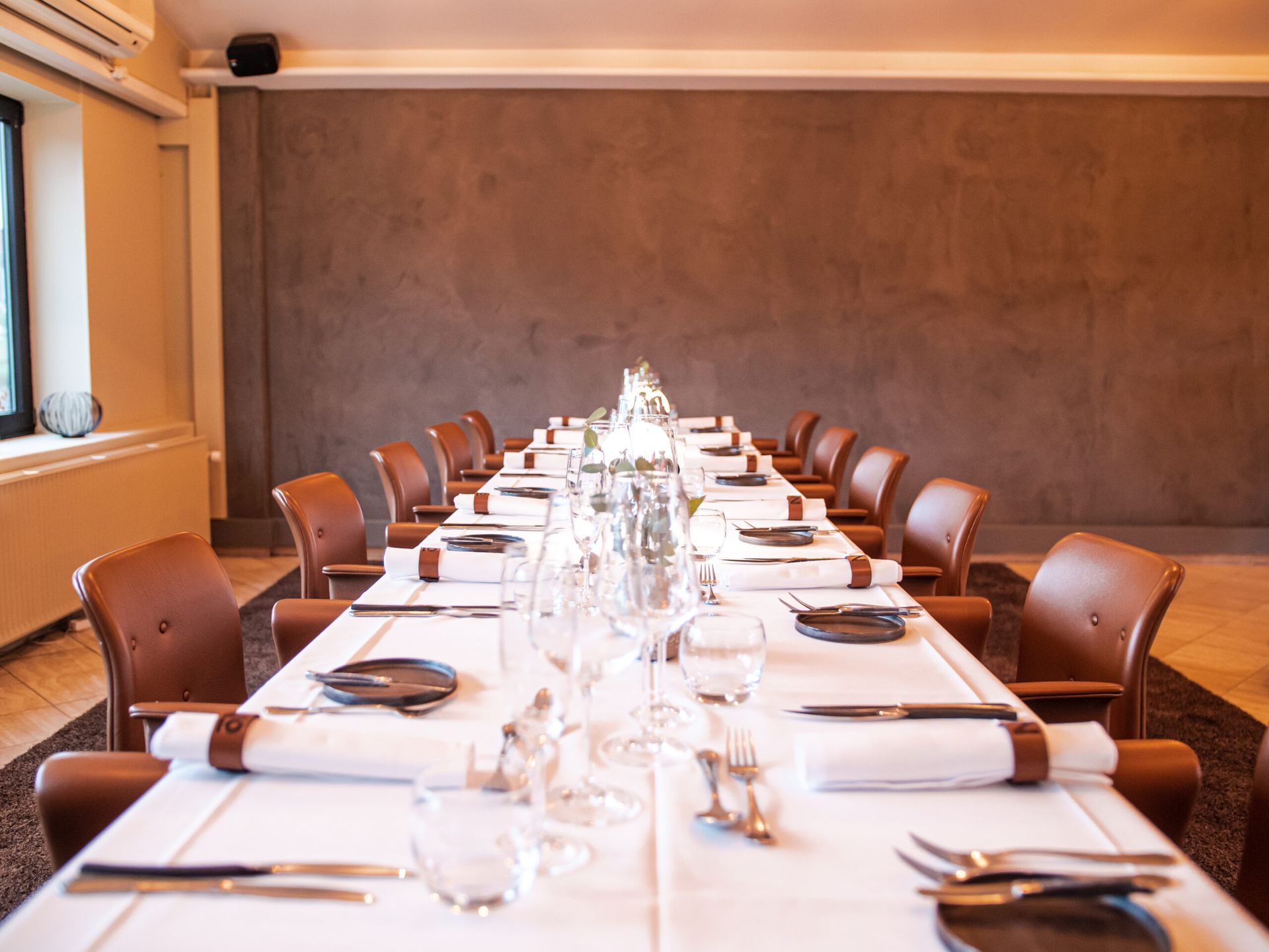 Long dining table set for a meal in a brown-walled room with brown leather chairs.