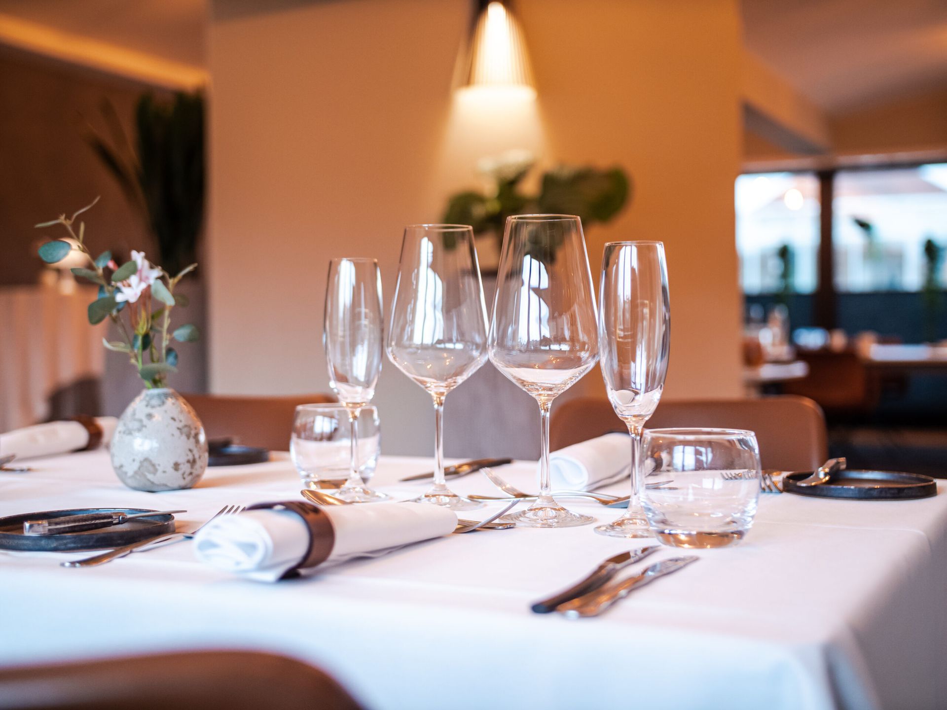 Table setting in a restaurant with wine glasses, silverware, napkins, and a small vase with flowers.