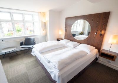 Hotel bedroom with double bed, large wooden headboard with circular mirror, window, armchair, and side table.
