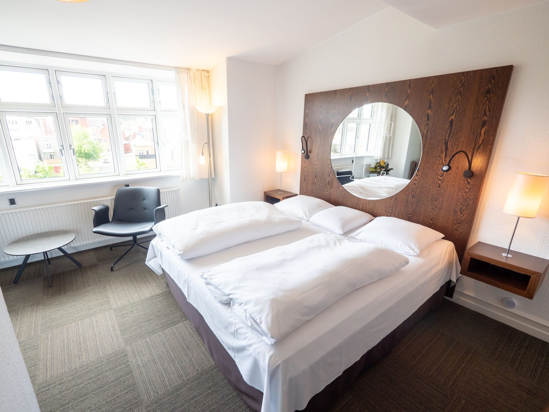 Hotel bedroom with double bed, mirror, window, chair, and side table.