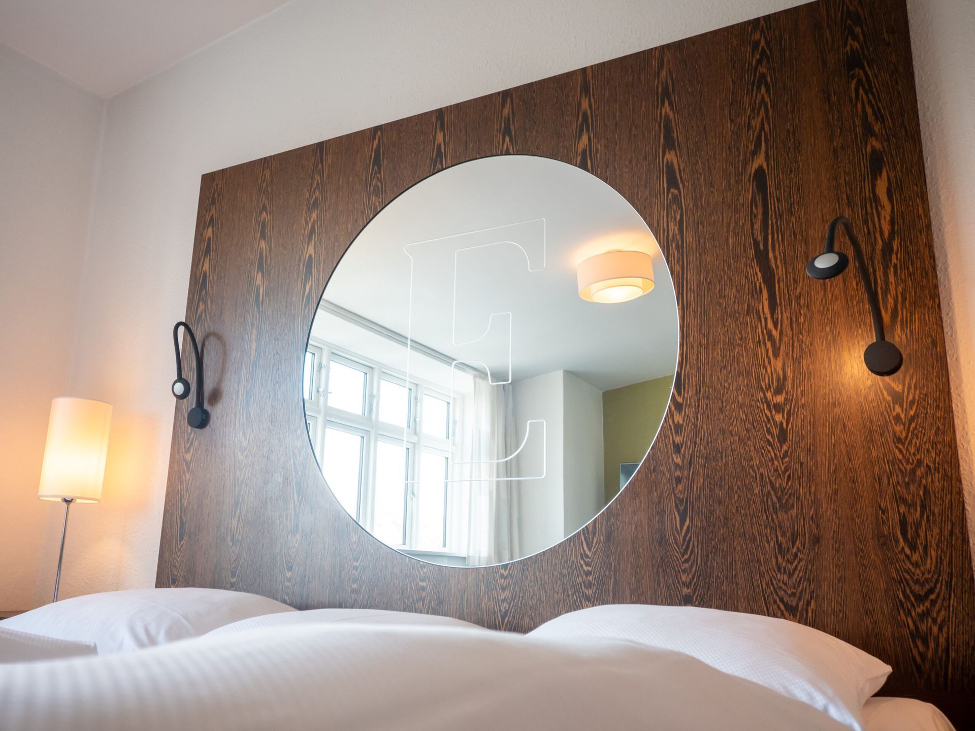 Bedroom with round mirror, wooden headboard, two lamps, and white bedding.