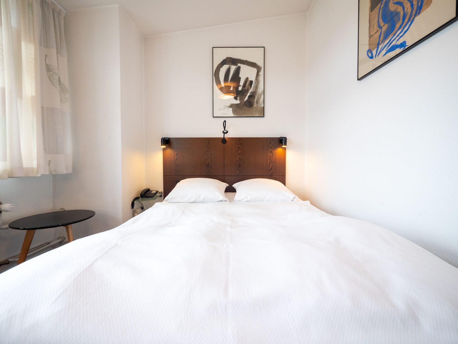 A bed in a white room with art above it, and a small side table on the left.