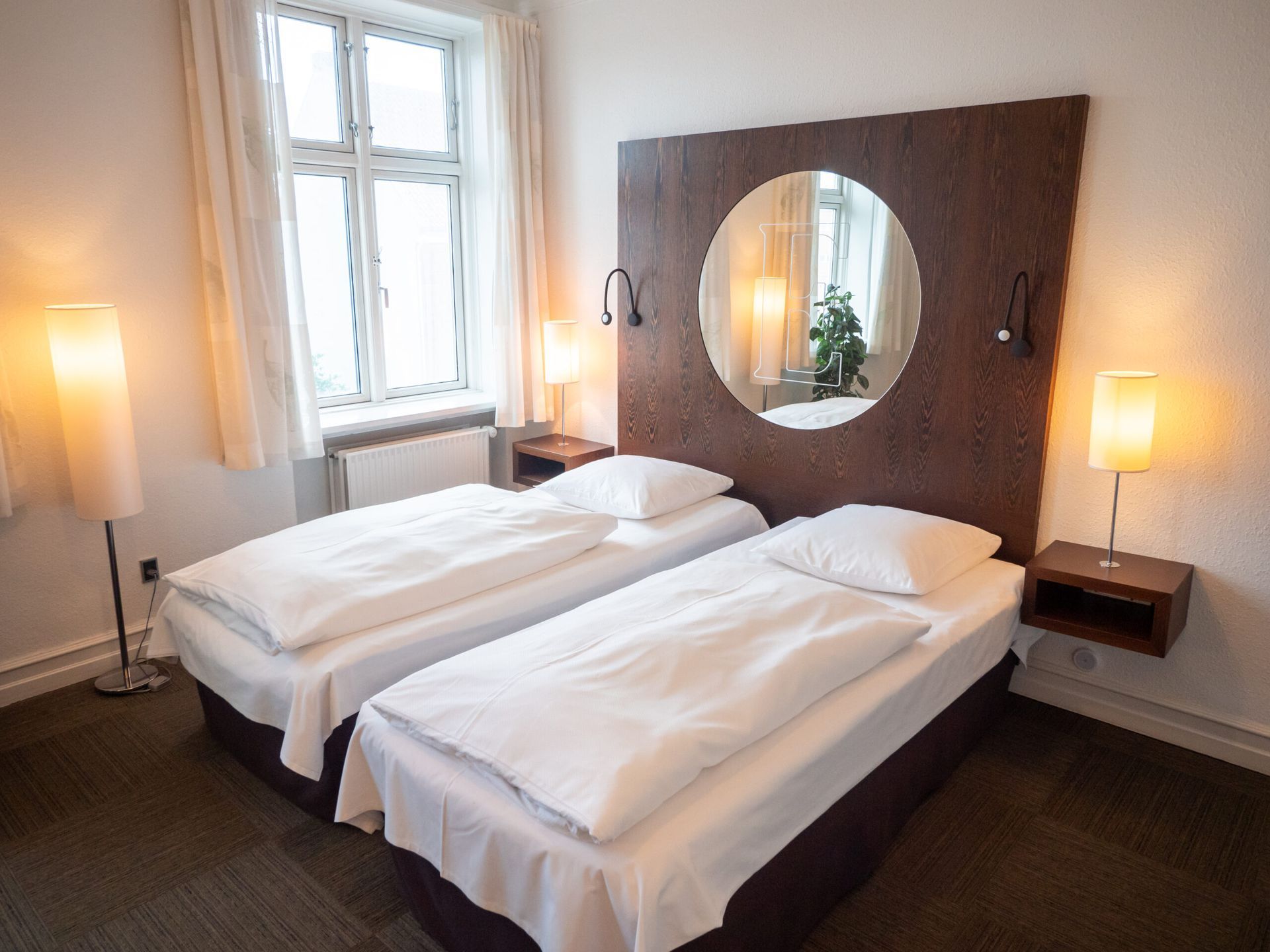 Twin beds in a hotel room with a large headboard, mirror, and lamps near a window.
