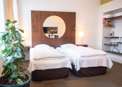 Hotel room with two twin beds, large round mirror, potted plant, and small shelves.