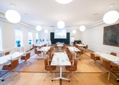 Meeting room with rectangular tables, brown chairs, white walls, and large round lights.