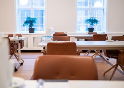 A light-filled conference room with brown chairs, white tables, and two potted plants near windows.