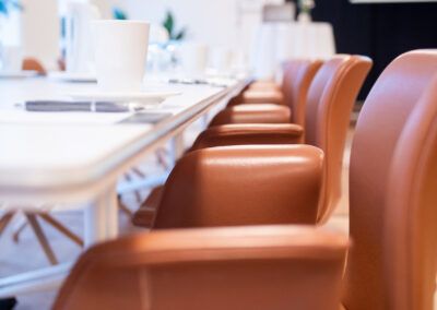Brown leather chairs line a long white table, set for a meeting in a bright room.