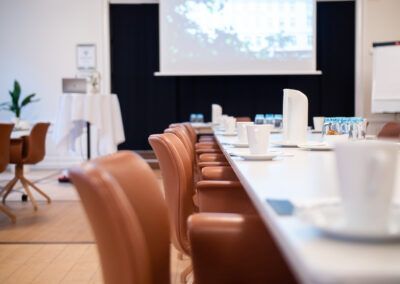 Conference room with brown leather chairs, long white table with cups, projector screen.
