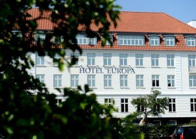 Hotel Europa building with white facade, red roof, and leafy foreground.