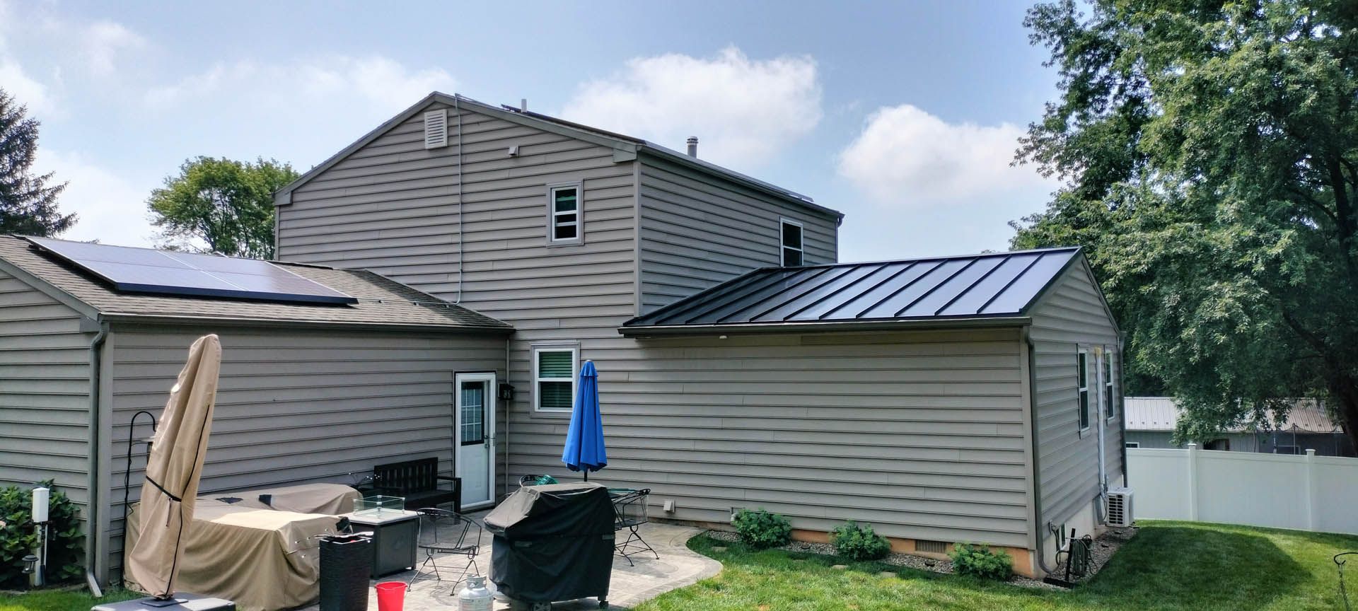 Backyard view of a house with solar panels and a metal roof, with a blue umbrella on a sunny day.