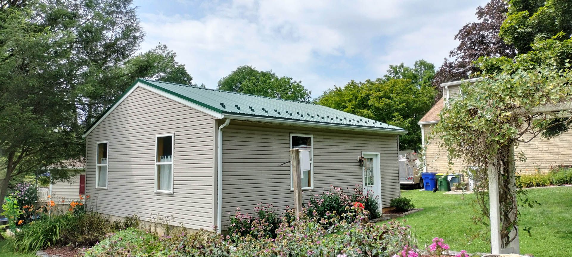 A small, light-colored building with a green metal roof sits in a yard with green grass and trees.