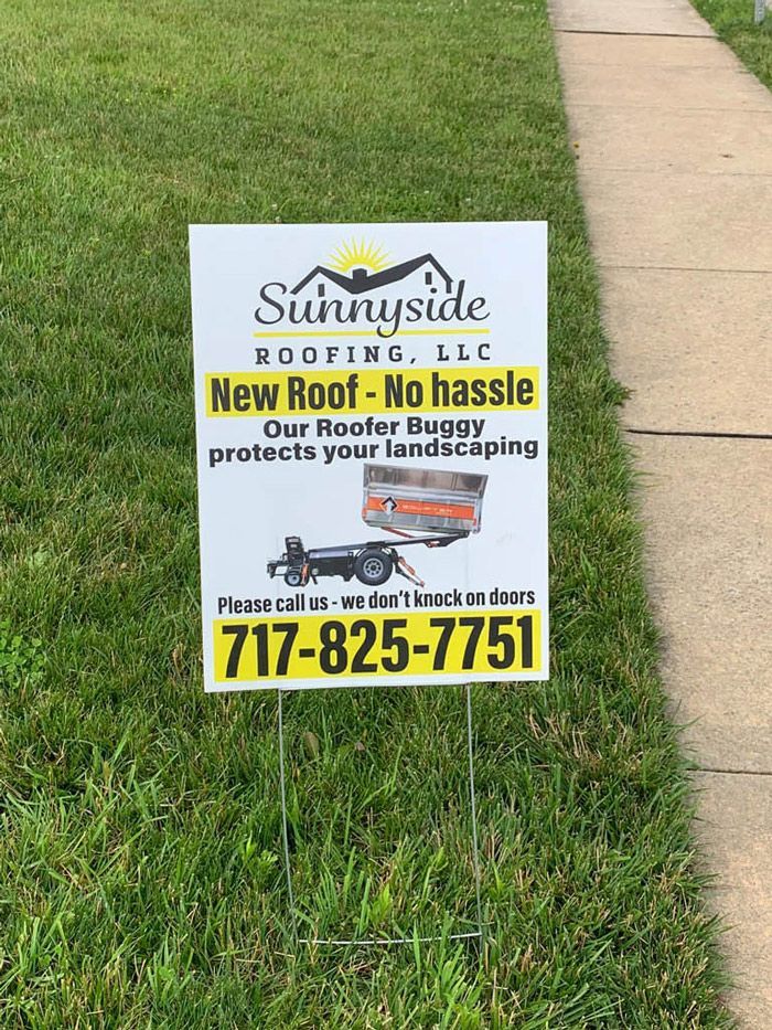 A sign for sunnyside roofing llc is sitting in the grass next to a sidewalk.