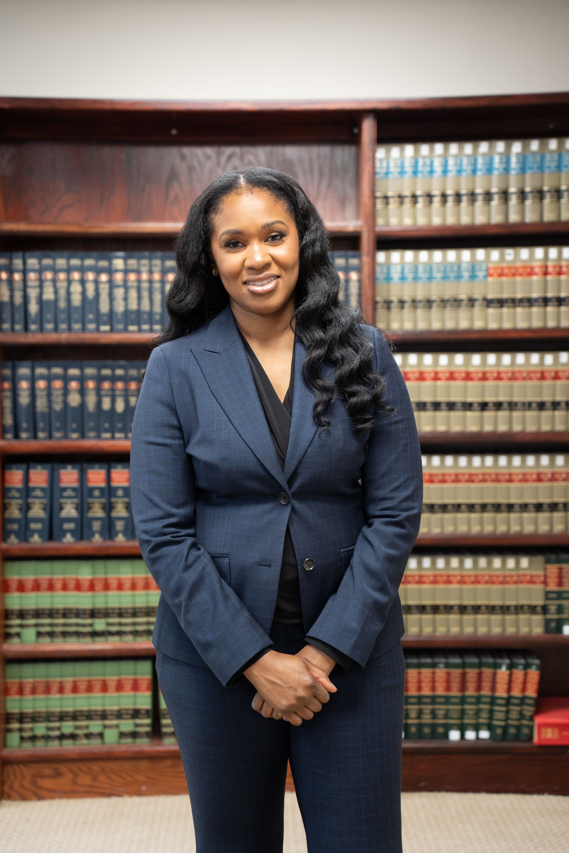 Woman in a blue suit stands in front of a bookcase. She has long, wavy hair and smiles slightly with her hands clasped in front of her.