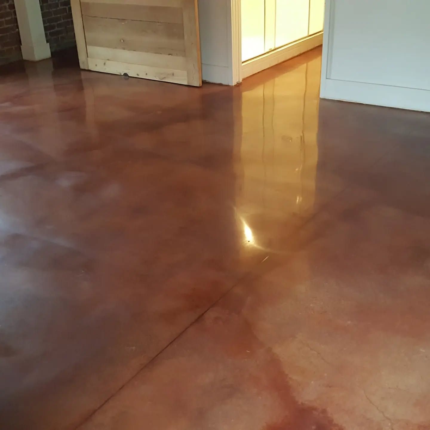 Polished reddish-brown concrete floor. Wooden door and white trim in the background. Sunlight reflects off the floor.