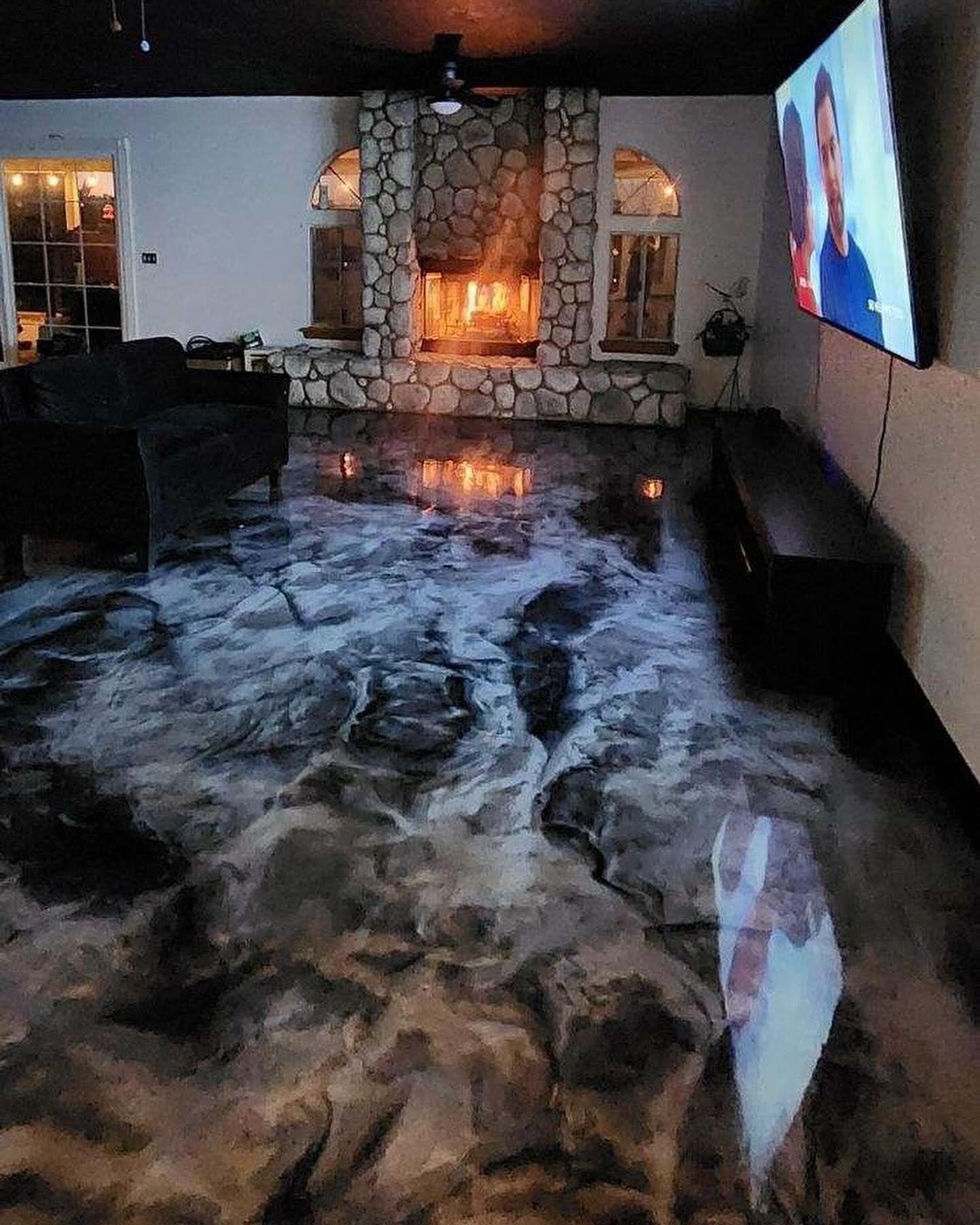 Living room with stone fireplace, black ceiling, and glossy gray epoxy floor reflecting firelight and TV screen.