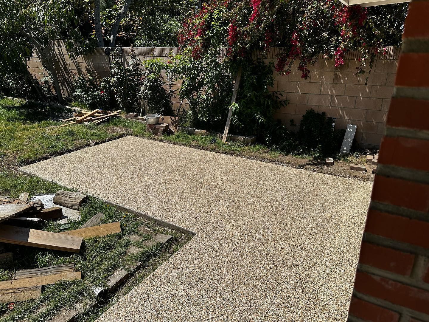 Gravel patio in a backyard with grass, surrounded by brick wall, plants, and construction materials.