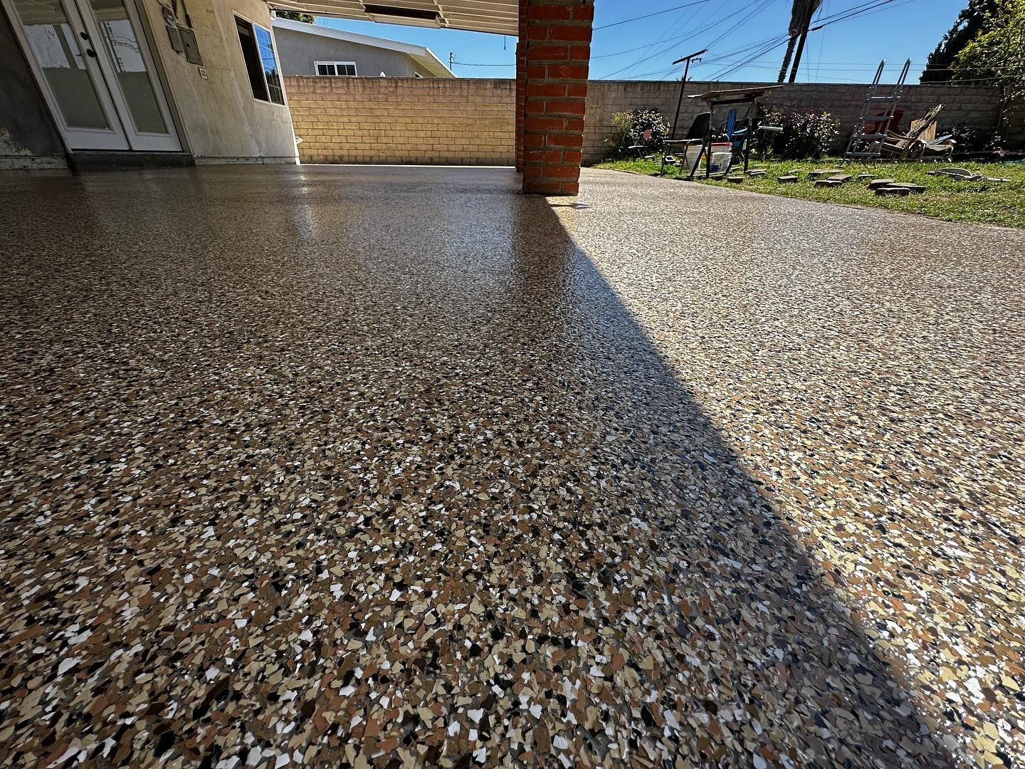 Epoxy-coated concrete patio with speckled brown, beige, and black flakes, under a brick-columned overhang; sunny outdoors.