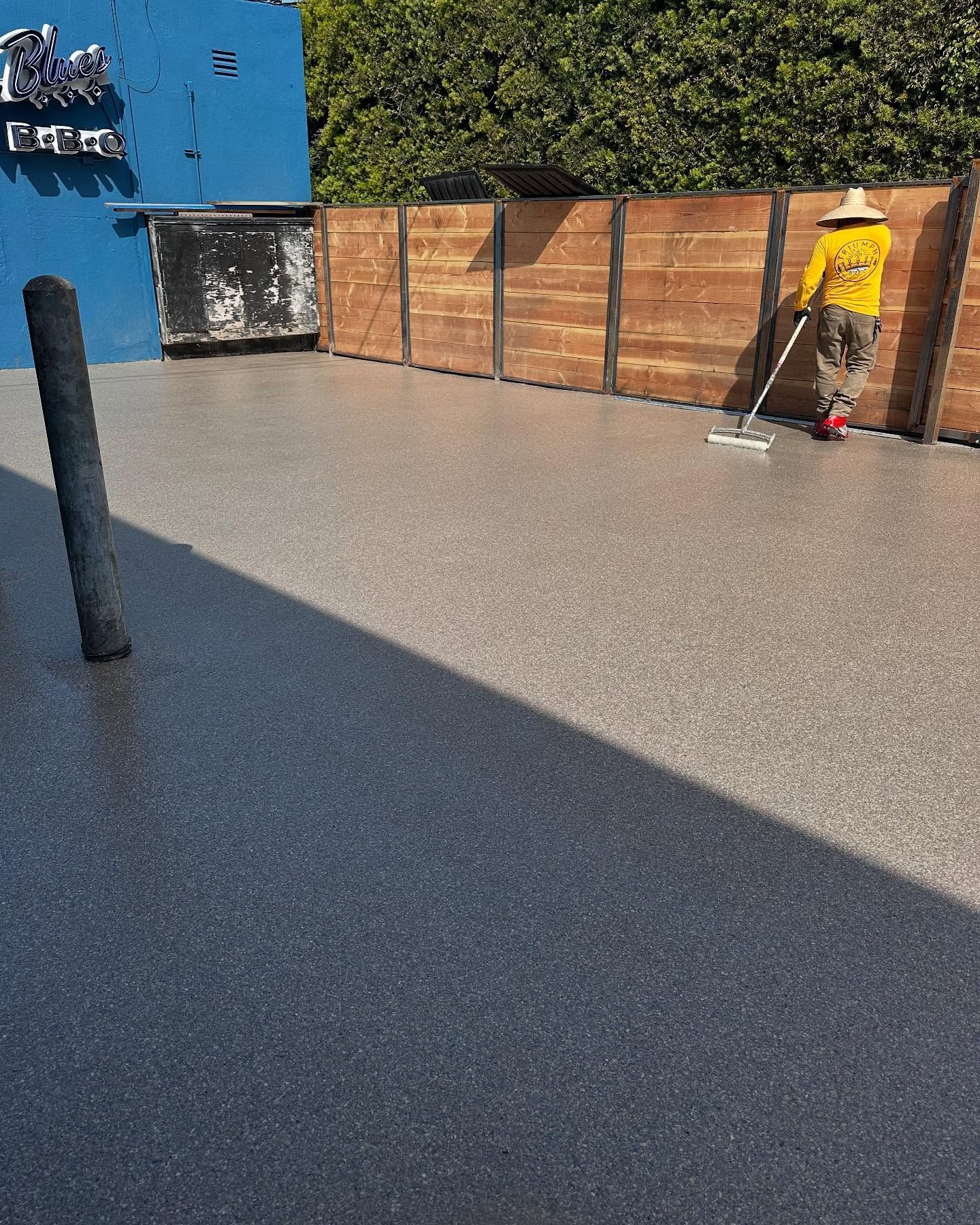 Person applying coating to a speckled gray surface with a long-handled tool, next to a wood fence.