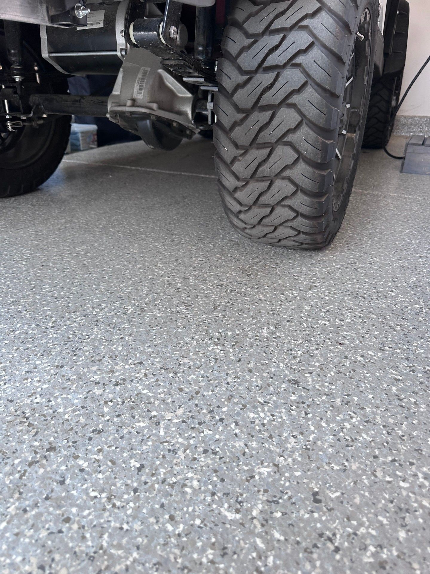 Close-up of a vehicle's tire on a speckled gray epoxy garage floor. The tire has a thick, patterned tread.