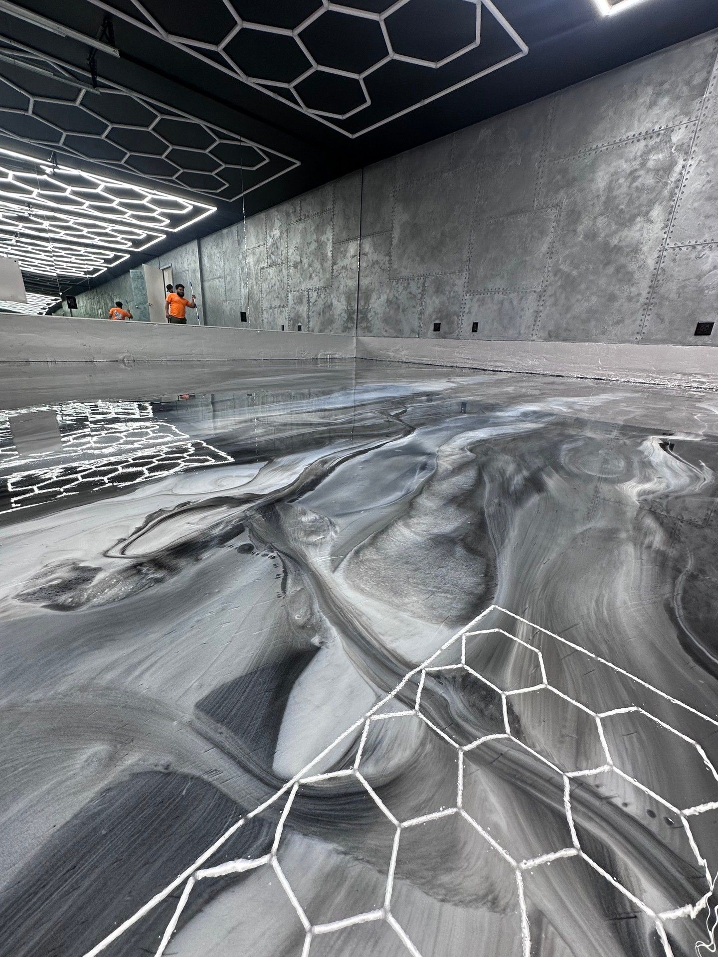 Black and white swirled paint on a floor, with honeycomb lighting. Gray walls and a person in the background.