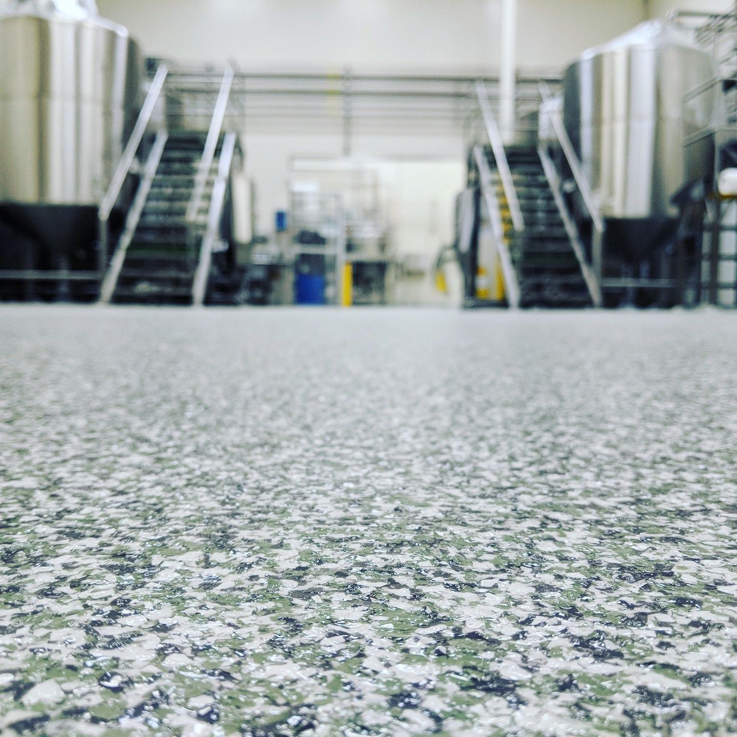 Close-up of a speckled floor in a brewery, with large stainless steel tanks and stairs in the blurred background.