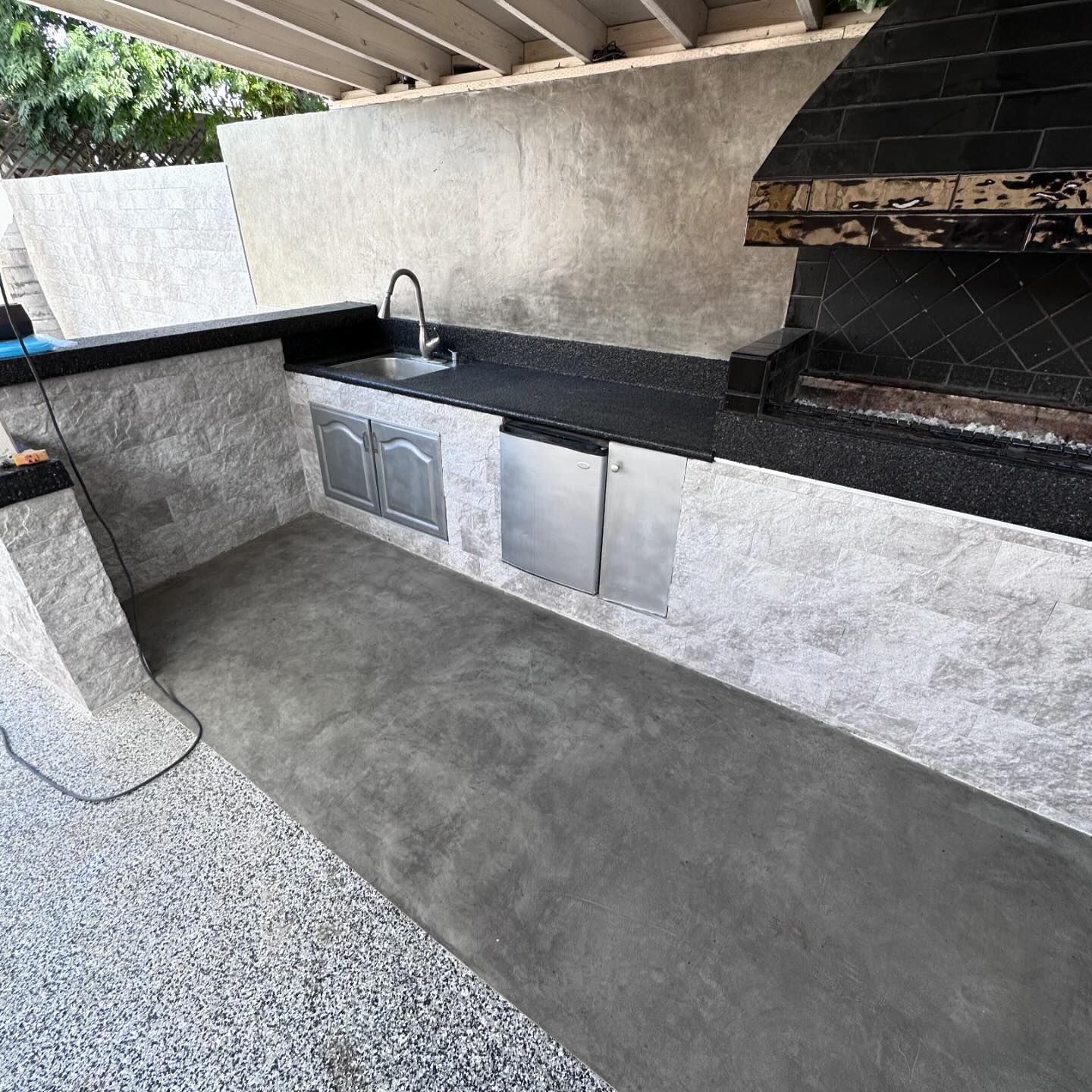 Outdoor kitchen with gray stone walls, black countertops, sink, stainless steel appliances, and built-in grill.