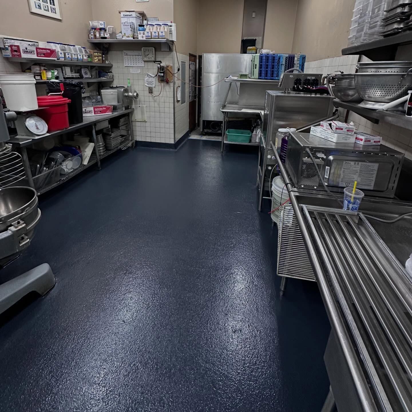 A commercial kitchen with stainless steel appliances, shelves with supplies, and dark blue flooring.