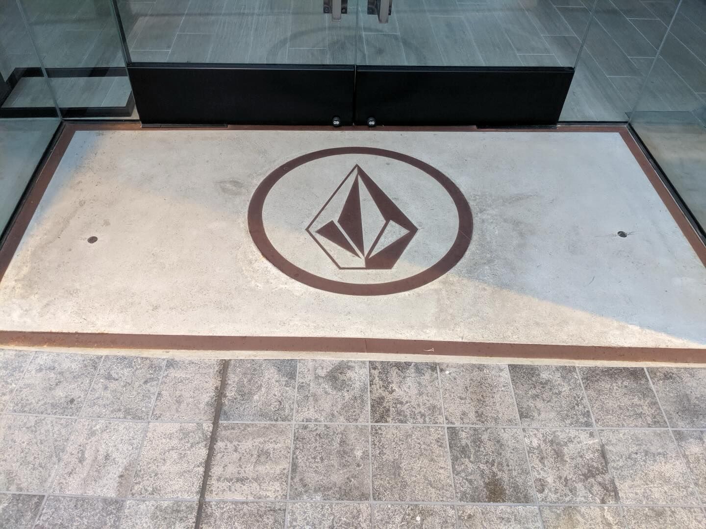Volcom logo embedded in a light-colored floor, framed by a dark brown border. Entrance with glass doors in background.