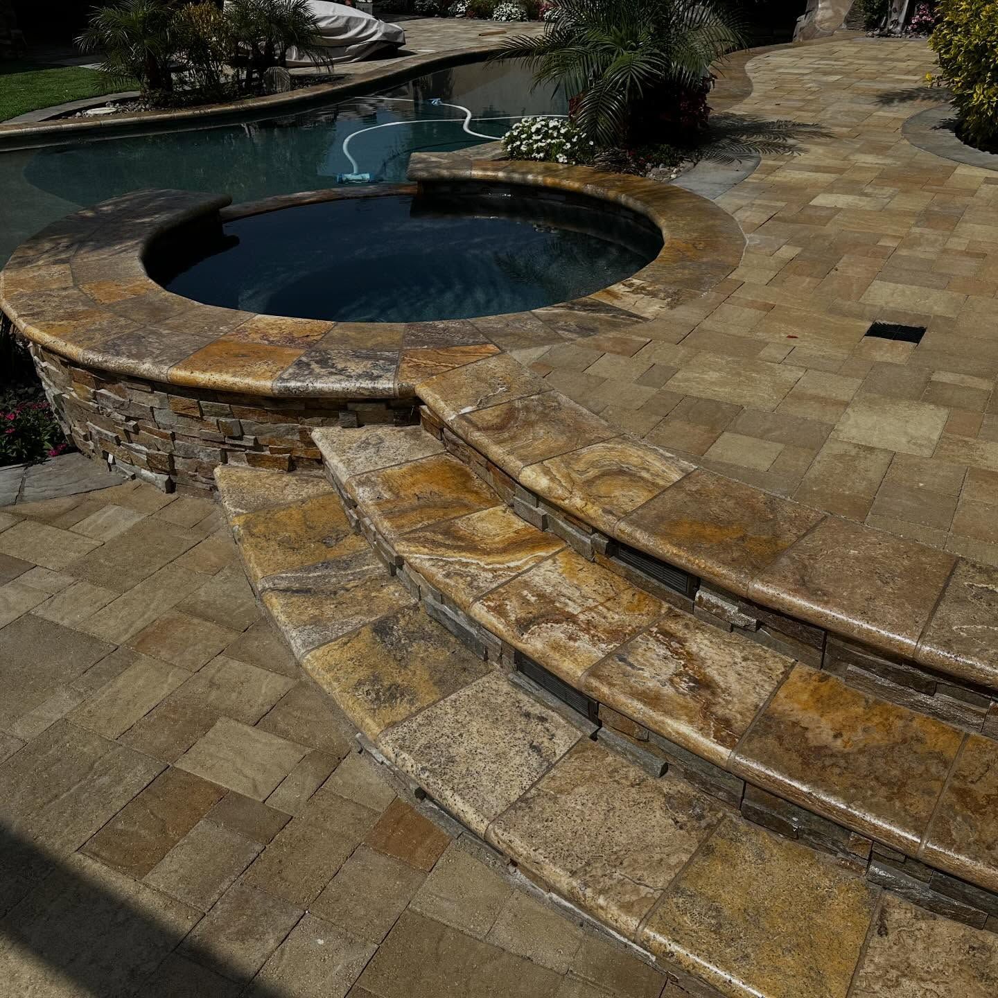 Stone patio with spa, pool, and steps. Tan and brown paving stones. Sunny outdoor setting.