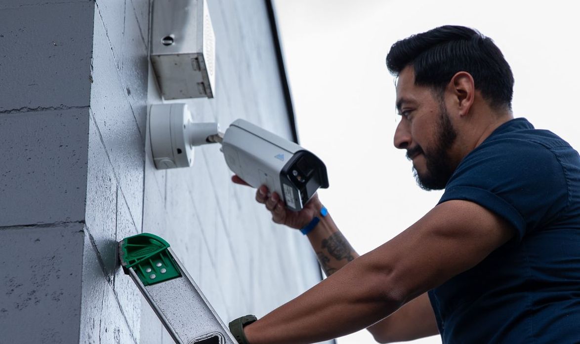 Man on ladder installing security camera on building.